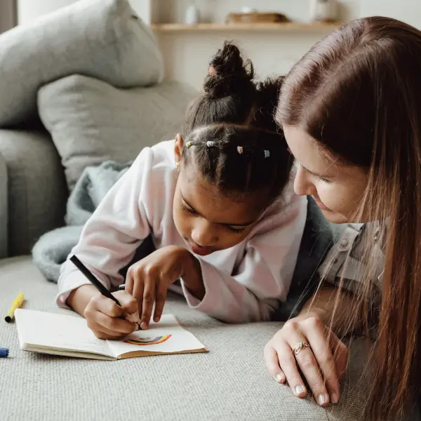 Foster care child drawing a rainbow on a card with the help of her foster care parent.